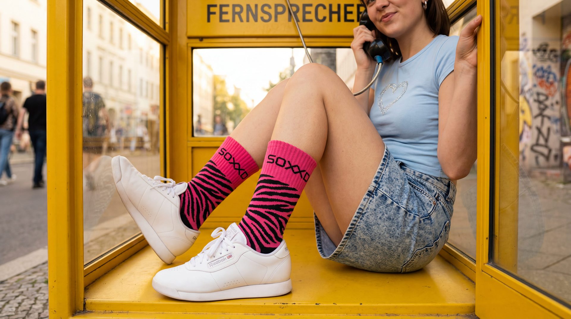 Friend on a yellow Berlin phone booth, pink-zebra SOXXD socks crossed up.